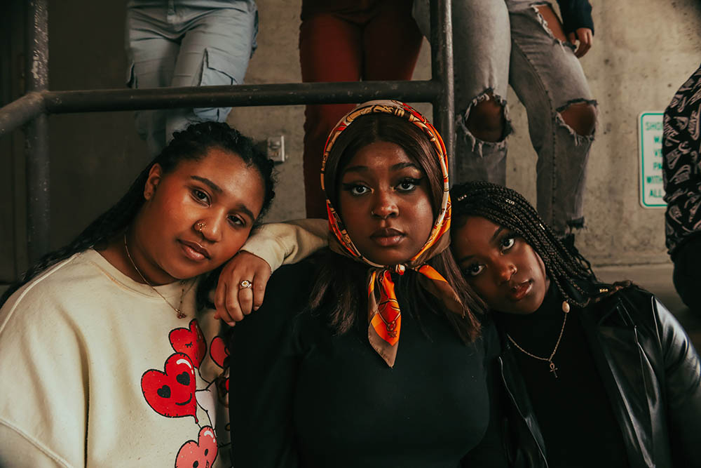 Three Black women leaning on each other's shoulders and looking into the camera with serious expressions