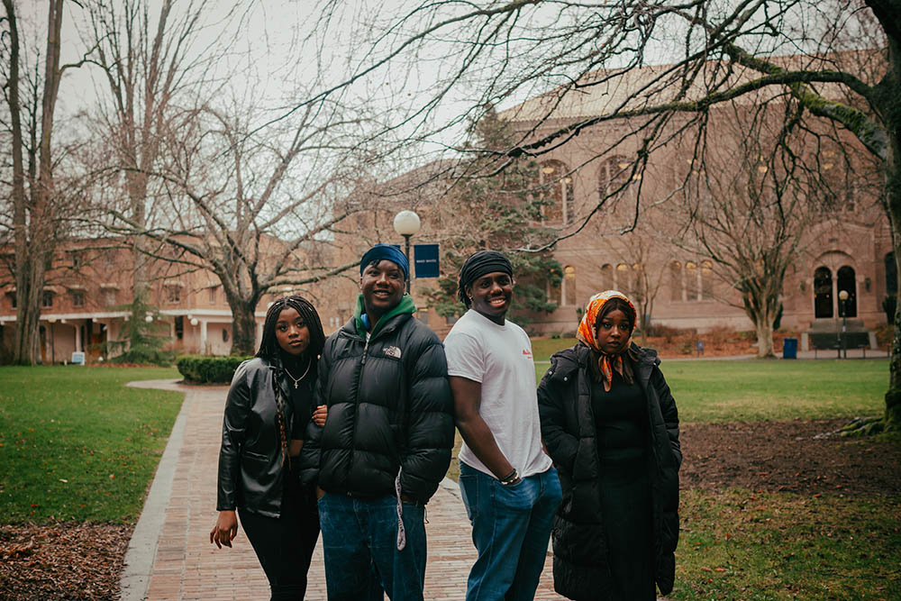 Four students from the Black Student Coalition pose together on a walkway in front of Old Main; the two men in the middle smile casually while the two women on either side of them strike a serious pose