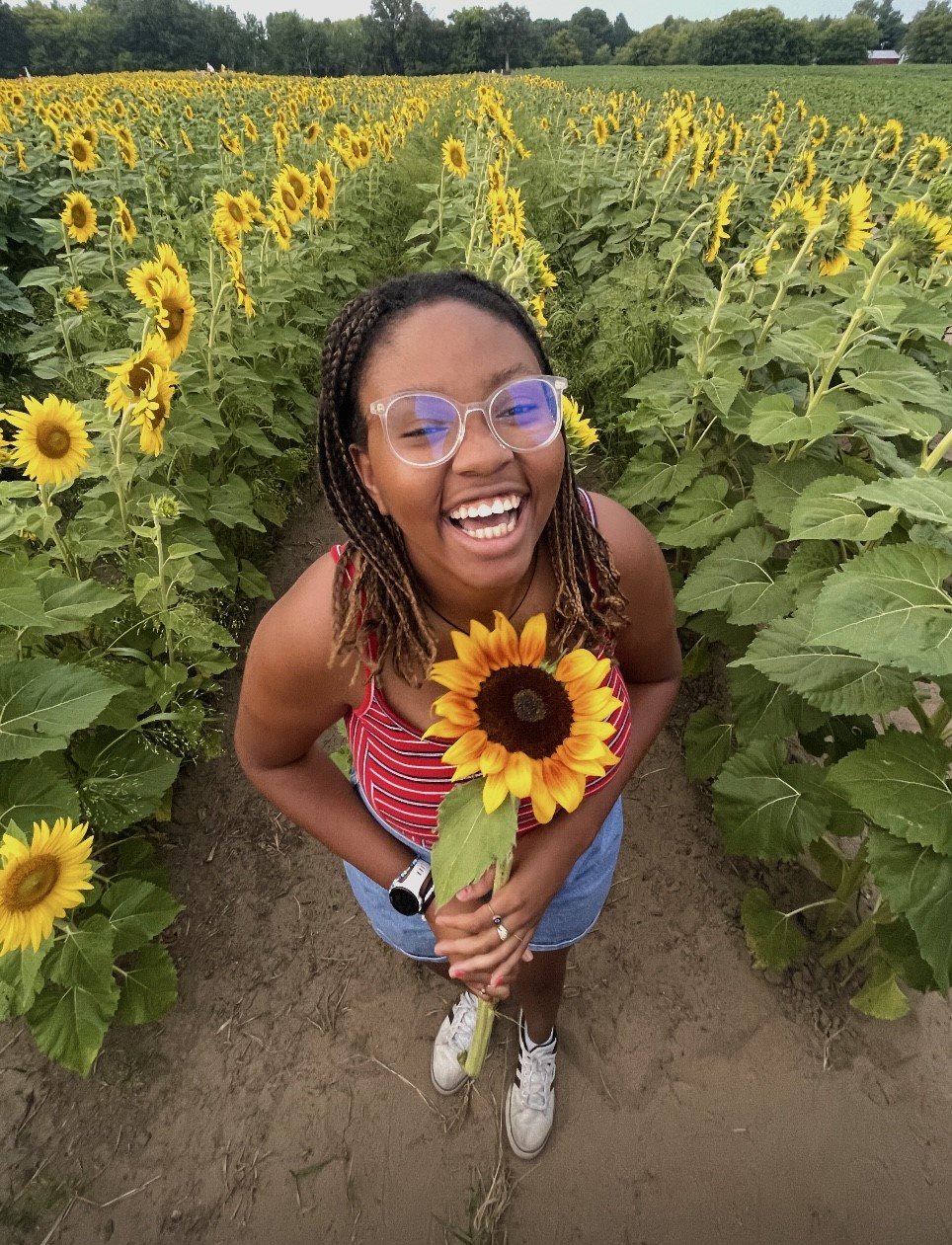 Faith stands smiling wide in an open field of sunflowers holding a single bright yellow and orange rimmed sunflower.