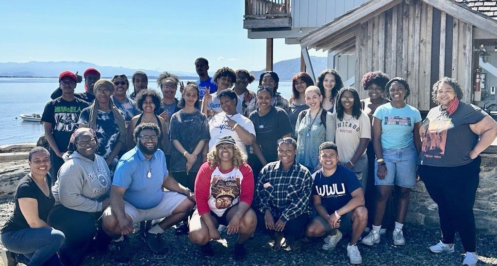 Group of Black Coalition Students smiling in the sunshine, wearing t-shirts and shorts in front of a wooden outbuilding perched on the edge of Bellingham Bay.